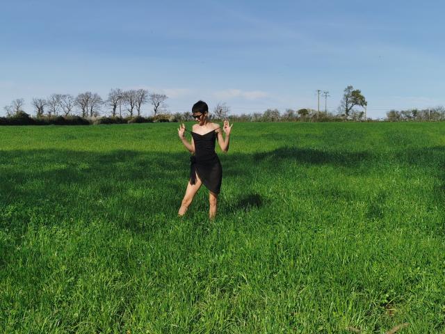LaPhoenixElo stands in a green field, wearing a black dress, raising her hands with fingers pointing up, her bare legs and black hair visible, under a blue sky.