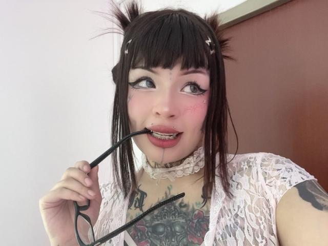 StarWitch with black pigtails, pink eyeshadow, tattoos, and a nose ring, biting black glasses, wearing a white lace top, and a choker. She's looking up with a playful expression.