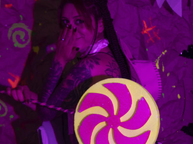 SofiFw has tattoos, covering her arms and neck, with dark nail polish, holding a pink and yellow swirl candy, lying under a pink blanket, in purple lighting.