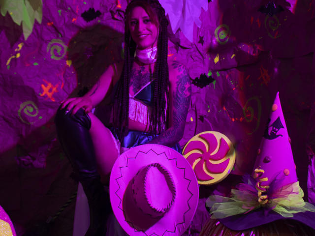 SofiFw with braided hair, purple and black outfit, thigh-high boots, sitting, smiling, surrounded by a hat, candy, and witch hat, in purple neon lighting, sexy, cute.