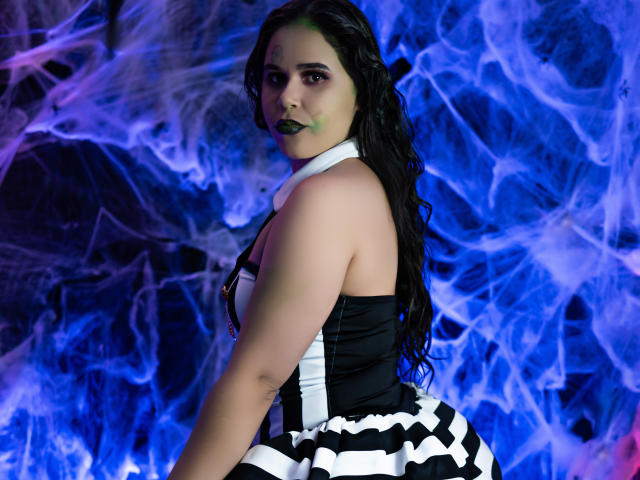 DanTaylor with dark wavy hair, black lipstick, white collar, black and white striped dress, facing camera, showing her ass, against blue and purple smoke.