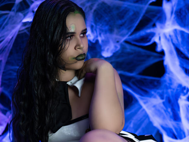 DanTaylor with dark wet hair, green face paint, black lipstick, and white black dress, crouching, holding knee, blue and purple smoke in background. Boobs, ass, tits, pussy not visible.