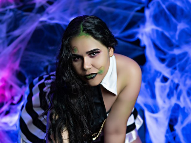 DanTaylor has dark green makeup on her face, black wavy hair, and black lipstick. She's wearing a white collar with a black and white striped dress, leaning forward.