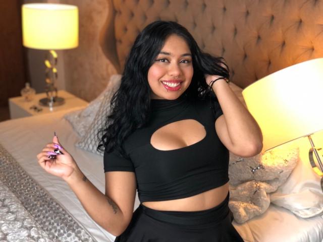 NicoleSmithy with black hair, smiling, wearing a cutout black top, holding a purple sex toy, sitting on a bed with a tufted headboard and lamp in the background.