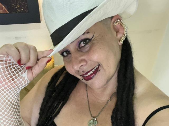 VampyBael with white hat, purple eyeshadow, black bra, fishnet glove, earrings, and nose ring, smiling, showing red nails, and wearing a necklace with a green pendant.