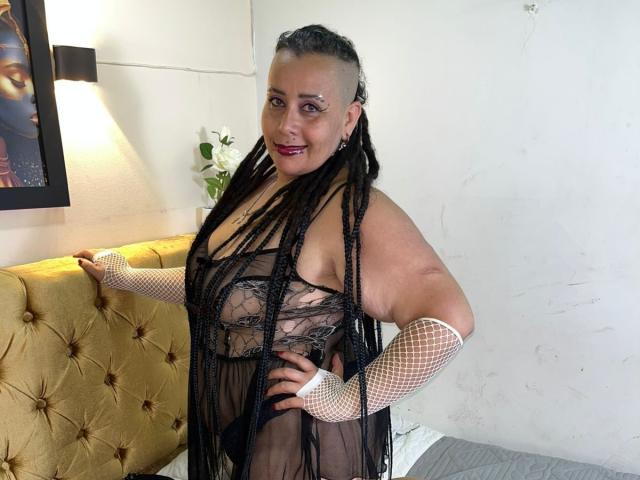 VampyBael with dark braids, sheer black lace dress, white fishnet gloves, and red lipstick, standing in a room with a yellow tufted headboard and a white flower.