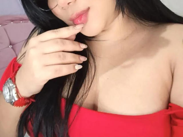 JessWintersLm with black hair, red top, and cleavage, biting her finger with pink lips, wearing glasses, and a silver bracelet on her right hand.