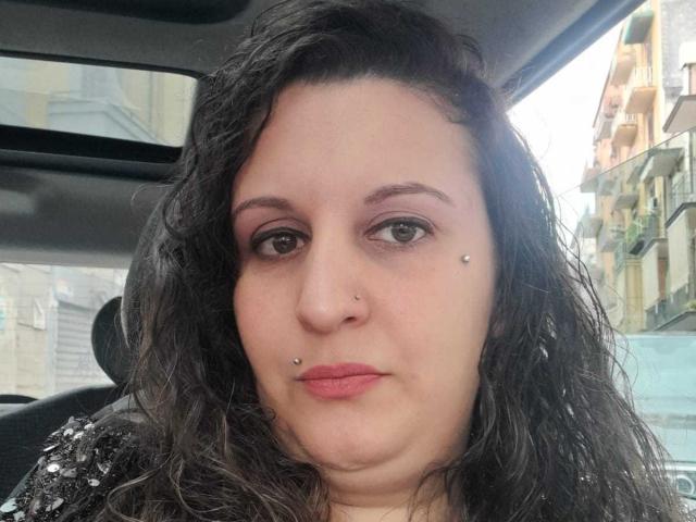 DelfinaSweet has curly black hair, brown eyes, pink eyeshadow, nose piercings, lip piercings, and a black sequined top, looking straight at the camera with a neutral expression.