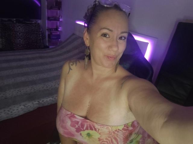 Vivien-Molinas, a bald woman with tan lines and a flower strapless top, showing off her boobs, tattoo on right shoulder, pierced nipple, purple lighting, and black chair.
