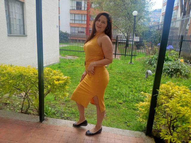 Karina Parker, con un sedere prosperoso, indossa un abito giallo a halter, scarpe basse nere, ed è in piedi su un patio in mattoni, con un giardino verde e cespugli gialli sullo sfondo, sorridendo alla macchina fotografica..