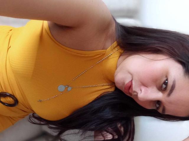 Karina Parker, com cabelo escuro, vestido amarelo texturizado e colar de prata, olhando para a câmera com um leve sorriso. Seios visíveis sob o vestido. Luz suave em seu rosto..