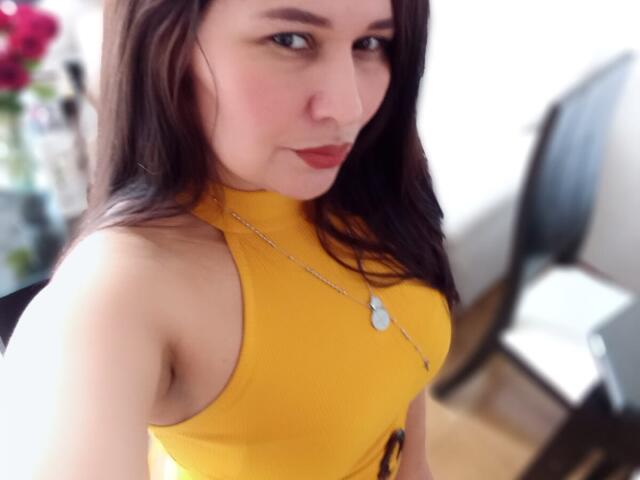 KarinaParker with long brown hair, yellow sleeveless top, red lipstick, small pendant necklace, and a slight smile, showing her bare shoulders and a hint of her boobs.