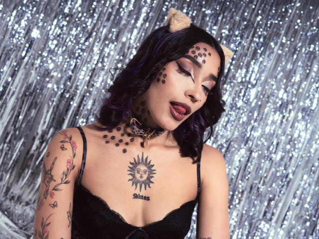 SalomeMille with dark purple hair, cat ears, black spots on face and chest, tongue out, black lace top, tattoo of sun face and "Suns" below, tattooed arm, silver backdrop.