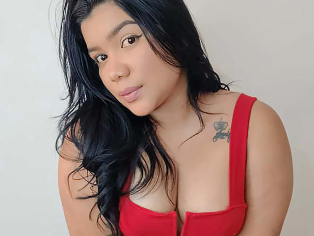 JessWintersLm with long black hair, wearing a red bra, showing her cleavage, has a small tattoo on her right shoulder, and is smiling softly.