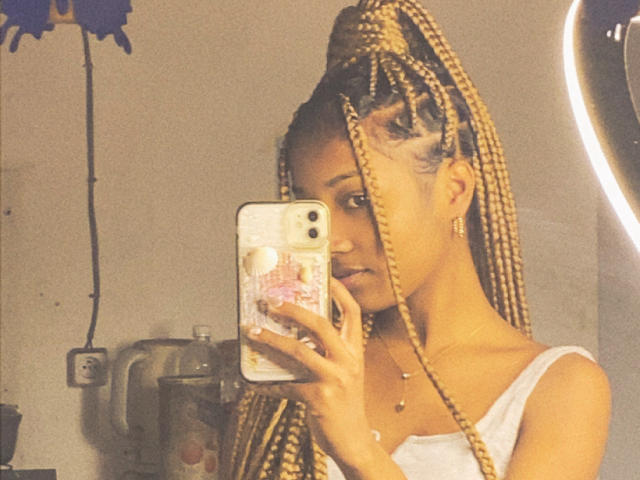 Portrait of FrenchQueen with long blonde braids, holding a phone in a mirror selfie. She wears a white tank top, small earrings, and a necklace with a small pendant. Her braids are thick and neatly arranged. The phone case has a floral design. The mirror has a circular, glowing edge. The background is blurry. Warm, slightly grainy lighting. Slight shadow on her face. Her expression is neutral. The image has a casual, everyday feel.