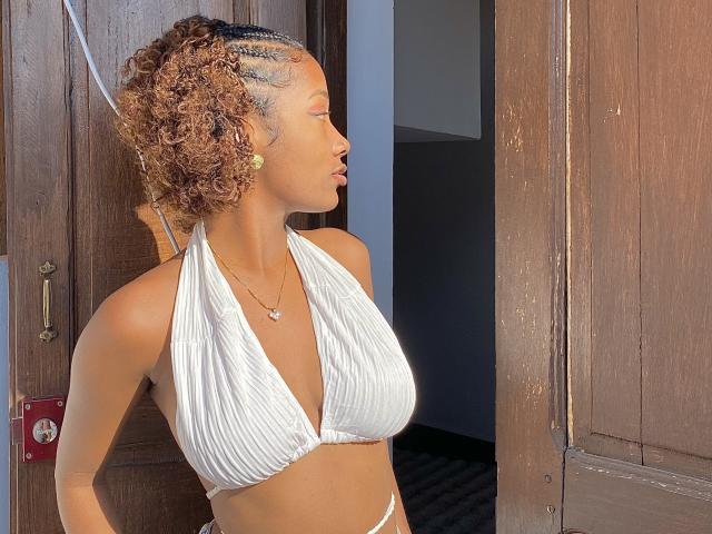 FrenchQueen with curly brown hair in a white halter bikini, showing her tits, standing in a doorway, looking to the side, wearing a necklace, in a sunny light.