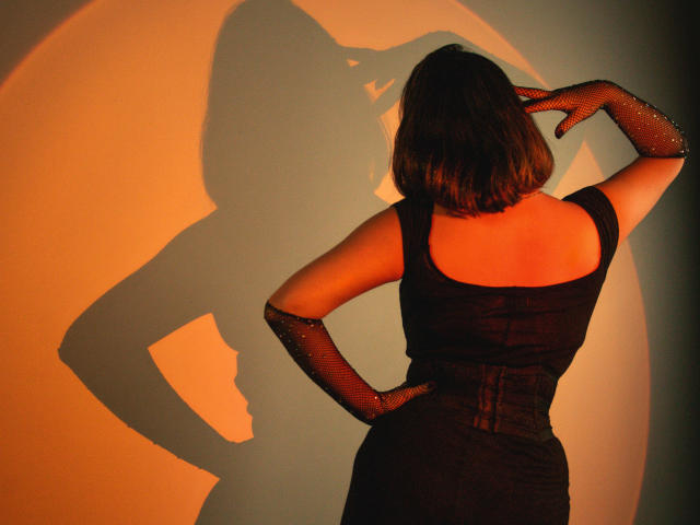 GoddessBar with back to camera, wearing black dress and sparkly fishnet gloves, hands on hips, shadow of her ass and tits on wall, warm orange light.
