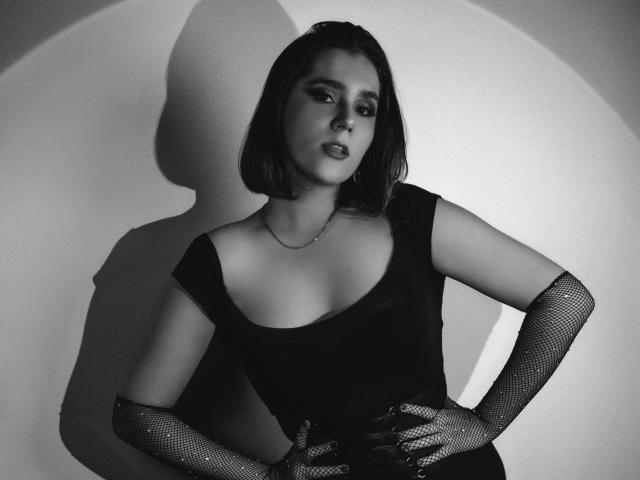 GoddessBar with short dark hair, wearing a black dress with a deep neckline, and fishnet gloves, hands on hips, looking seductive, shadowed in a black-and-white photo.