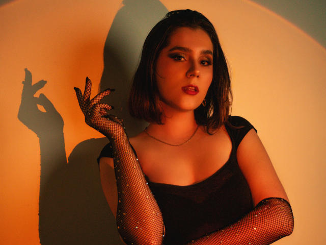 GoddessBar with short brown hair, dark makeup, red lips, wearing black fishnet gloves and dress, hand up, shadow on wall, warm light, looking at camera, showing off her tits.