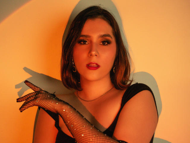 GoddessBar with shoulder-length brown hair, red lipstick, black fishnet gloves, and a black top, looking seductively at the camera with a slight smile. Warm orange lighting.