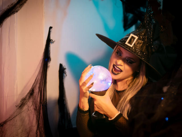 ChantalSex69 with a witch hat, holding a glowing orb, has dark lipstick and blonde hair, wearing a black top, with spooky black webbing in the background.
