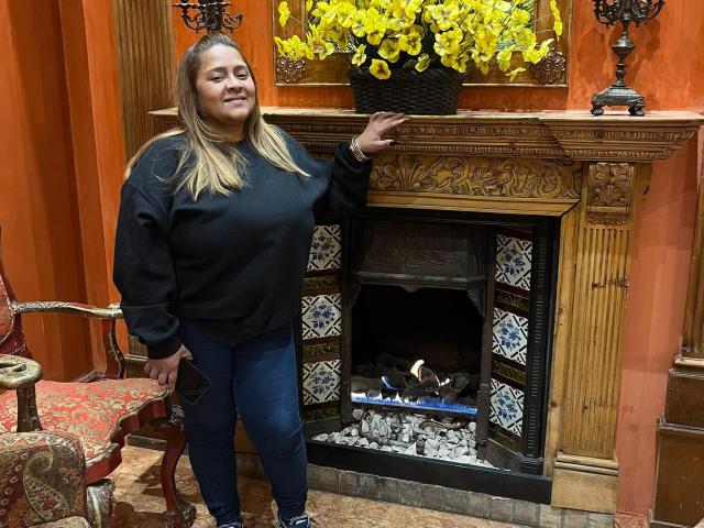 MonzerratBoyer stands in front of a wooden fireplace, wearing a black sweatshirt and blue jeans, with blonde hair, smiling, and resting her arm on the ornate fireplace mantle.