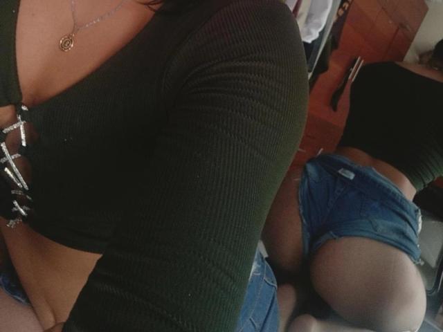 YennettSphinx in a black ribbed top with cleavage, wearing denim shorts, showing her ass in a mirror reflection. She's kneeling, highlighting her butt and panty-line.