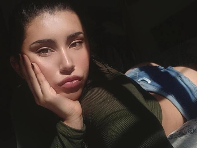 YennettSphinx lying on her side, hand on cheek, green top, denim shorts, sunlight on face, brown eyes, slightly parted lips, dark background, subtle makeup, dark hair, soft shadows.