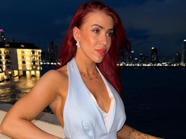 JustSophia-hot with bright red hair, wearing a light blue halter top with deep V-neck, stands against a nighttime waterfront. She has pink earrings, a nose ring, and tattoos on her right arm. Her makeup includes bold eyeliner and pink lipstick. The city lights in the background are blurred, highlighting her in the foreground. She's looking to the right with a slight smile. The image has a sharp, clear focus on her.