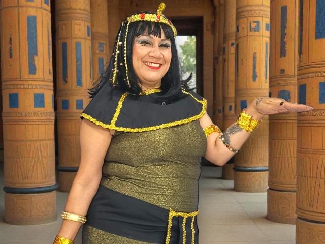 MatureYayita with dark hair, gold and black outfit, Egyptian headpiece, gold jewelry, smiling, holding hand out, showing tattoo on left arm, standing in front of wooden columns.