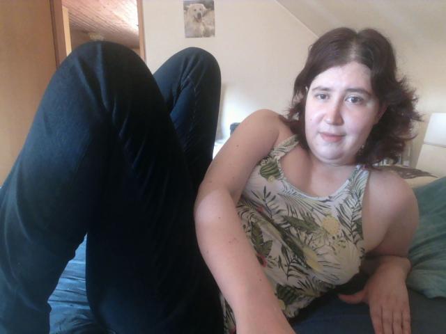 A photograph of SexyLucy69, a plus-sized woman with fair skin and shoulder-length brown hair, lying on a bed. She wears a sleeveless, floral-patterned top and black pants. Her legs are raised and bent at the knees, with her feet pointed upward. She has a slight smile and looks directly at the camera. The bed has a blue sheet, and a light blue pillow is visible. A small wall art piece is in the background. The room has a wooden door and a lamp on the right.
