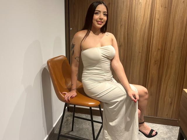 BellaVibes, with long dark hair and tattoos, sits on a tan chair, wearing a strapless white dress, showing her tits and smiling, with her ass barely visible.