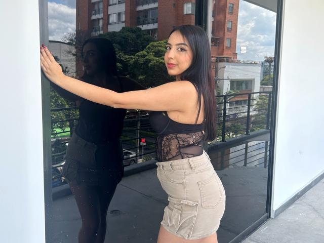 BellaVibes è in piedi in una porta, indossa una maglia di pizzo nero e pantaloncini beige, mostrando il sedere e riflettendosi nello specchio. Sta guardando indietro con un leggero sorriso..