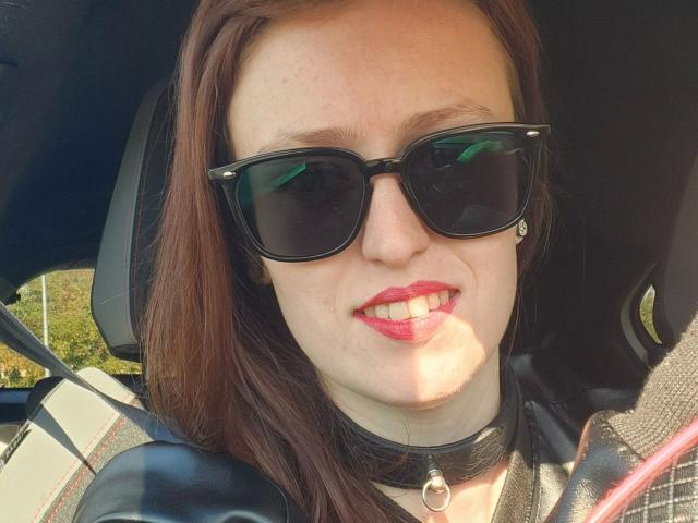 AvaNoir-hot with red lipstick, black sunglasses, and a black choker, smiling in a car, has brown hair and is wearing a black leather jacket.