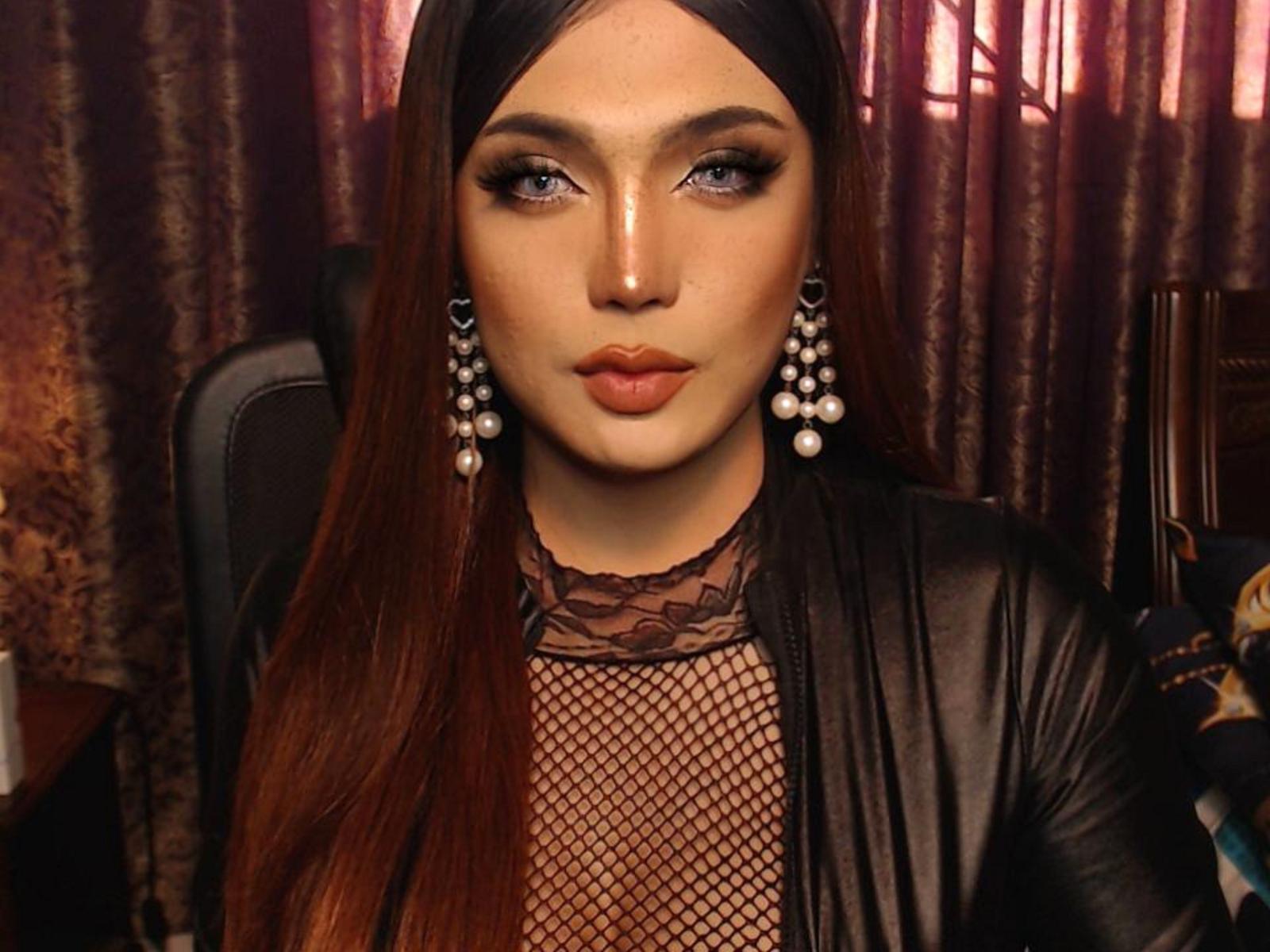 SchonBella has long, straight, auburn hair, wearing a black leather jacket over a mesh top. She has large, dark eyes with heavy black eyeliner, full, orange-red lips, and wears large, pearl-studded earrings. Her makeup is bold and dramatic. The background has dark, textured curtains. The lighting is warm, highlighting her facial features and hair. She looks directly at the camera with a neutral expression. The image has a high-contrast, glamorous style.