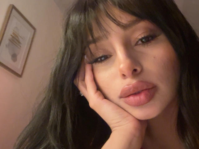 ThaliaCollins with dark, wavy hair and bangs, hand on cheek, full pink lips, slightly parted, looking at camera, brown eyes, soft lighting, subtle makeup, slightly tanned skin.