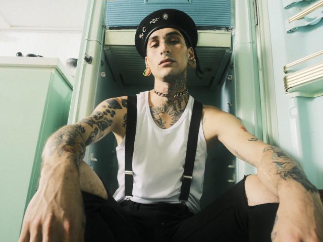 TonyoHatsumoto is a tattooed, slim young man with pale skin and dark hair, wearing a black beret with a star pattern, gold ear gauges, and a choker. He's in a white tank top with black suspenders and black pants, sitting with legs apart. He has tattoos on his arms and chest. The photo has a low-angle view, emphasizing his tattoos and relaxed posture. The background is a mint-green interior with a door and fluorescent lights. The image has a gritty, urban vibe with clear, sharp details.