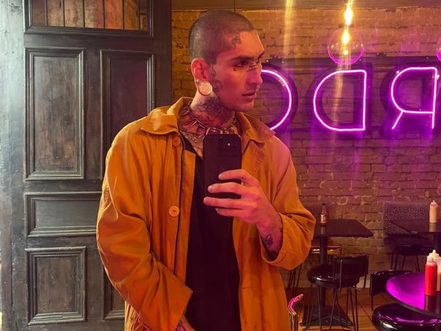 TonyoHatsumoto with shaved head, tattoos on neck and ear, wearing mustard coat, black shirt, holding phone, purple neon sign in background, standing by dark door, looking at mirror.