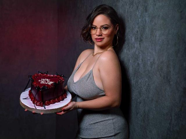 TaylorSexiest with big boobs, wearing a grey dress, holding a red chocolate cake with black decorations, standing against a dark wall, wearing glasses and a gold necklace.