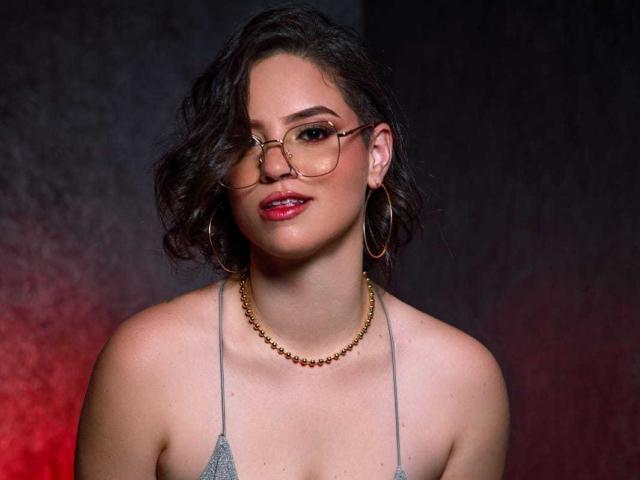 TaylorSexiest with wavy dark hair, glasses, red lipstick, gold hoop earrings, necklace, gray halter top, showing off her boobs, against a dark, textured background.