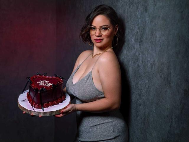 TaylorSexiest with glasses, wearing a gray dress showing big boobs, holding a red chocolate cake with cherries and black decorations, standing against a dark wall.