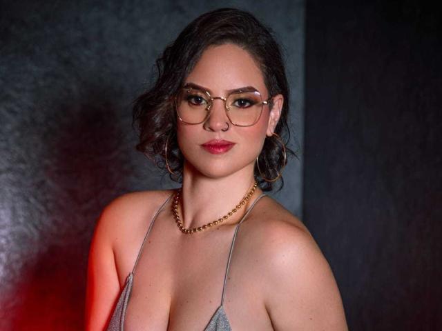 TaylorSexiest with curly brown hair, glasses, red lipstick, hoop earrings, gold necklace, and a grey, strappy top, showing her tits and a serious expression.
