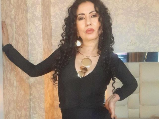 SamanthaAntolin stands confidently, hand on a lace curtain. She has long, curly black hair and wears a black long-sleeve top. A large, circular metallic necklace with multiple discs hangs from her neck. She has light skin, dark eyebrows, and wears bold red lipstick. Her right hand rests on her hip. The background features a beige, floral-patterned wallpaper and a reflective surface on the right. Her expression is neutral, lips slightly pursed. The image has a casual, slightly blurred quality.