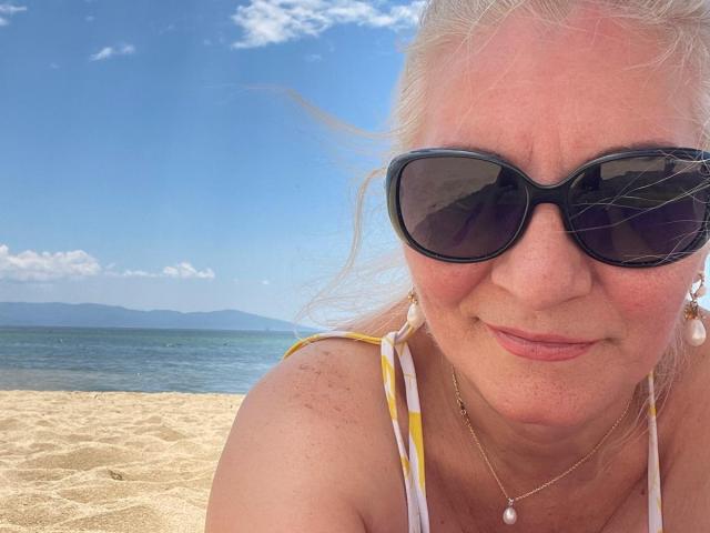 LadyJanyne with blonde hair, wearing black sunglasses, yellow-and-white striped bikini top, gold necklace, and subtle smile, on a sunny beach with clear blue water and distant mountains.