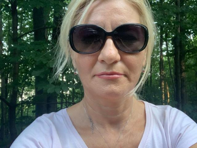 LadyJanyne with blonde hair, wearing black sunglasses, a white tee, and a delicate necklace, has a neutral expression, standing in a forested area, showing her nose and lips.