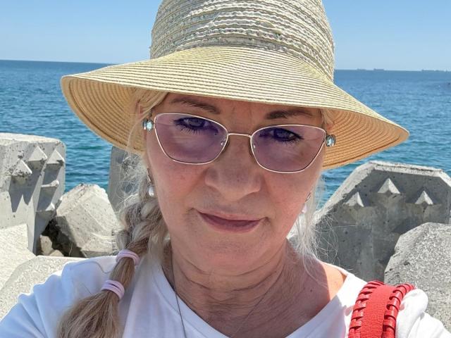 LadyJanyne with a straw hat, purple-tinted sunglasses, white shirt, and red bag, standing by the ocean, showing her ass-aging face and a subtle smile.