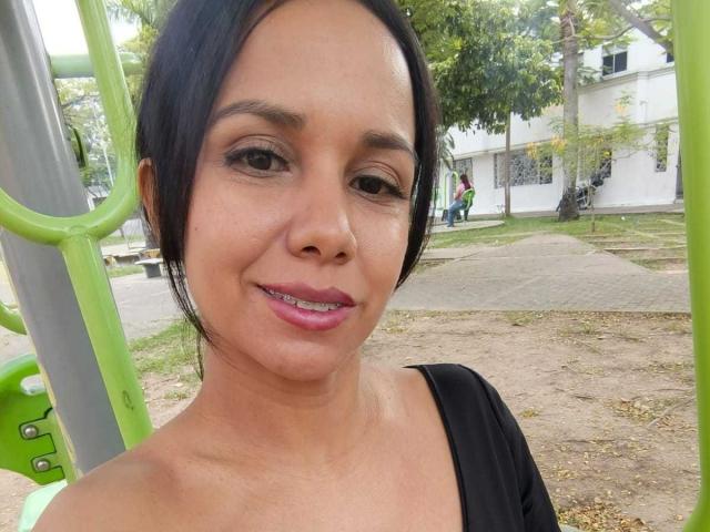 EvelinFoster has tan skin, black hair, brown eyes, and pink lipstick, smiling with her teeth showing, wearing a black top, standing near green playground equipment.