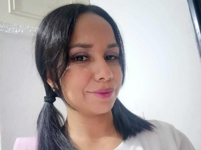EvelinFoster has dark brown pigtails, medium brown skin, pink lipstick, and a slight smile. She wears a white shirt and has small, subtle earrings.