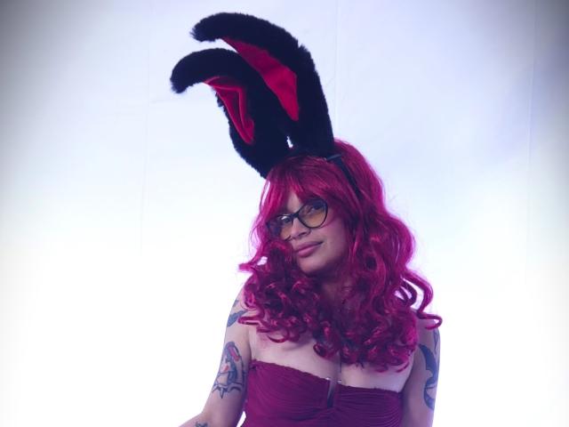 KoraSweet with bright pink curly hair, black fuzzy bunny ears, glasses, strapless red top, tattoos on her arms, smiling at the camera. Boobs visible, looking cute.