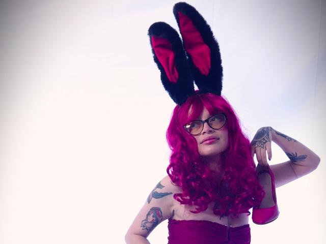 KoraSweet with pink curly hair, black and red bunny ears, glasses, and tattoos, wearing a purple strapless top, showing a hand with a ring.